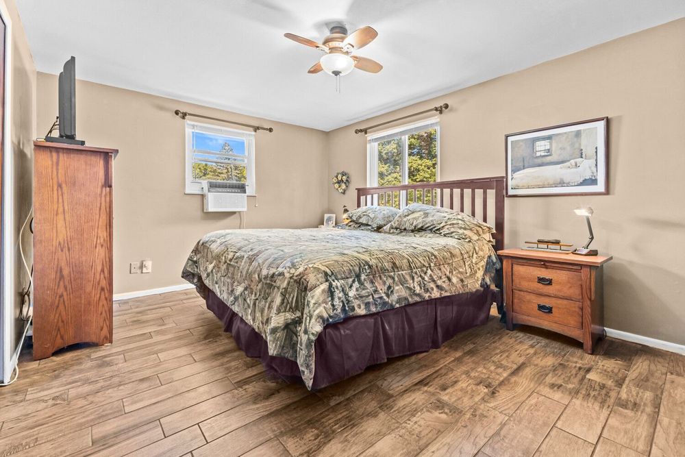 Spacious bedroom with hardwood floors, ceiling fan, two windows, and warm wood furniture pieces.