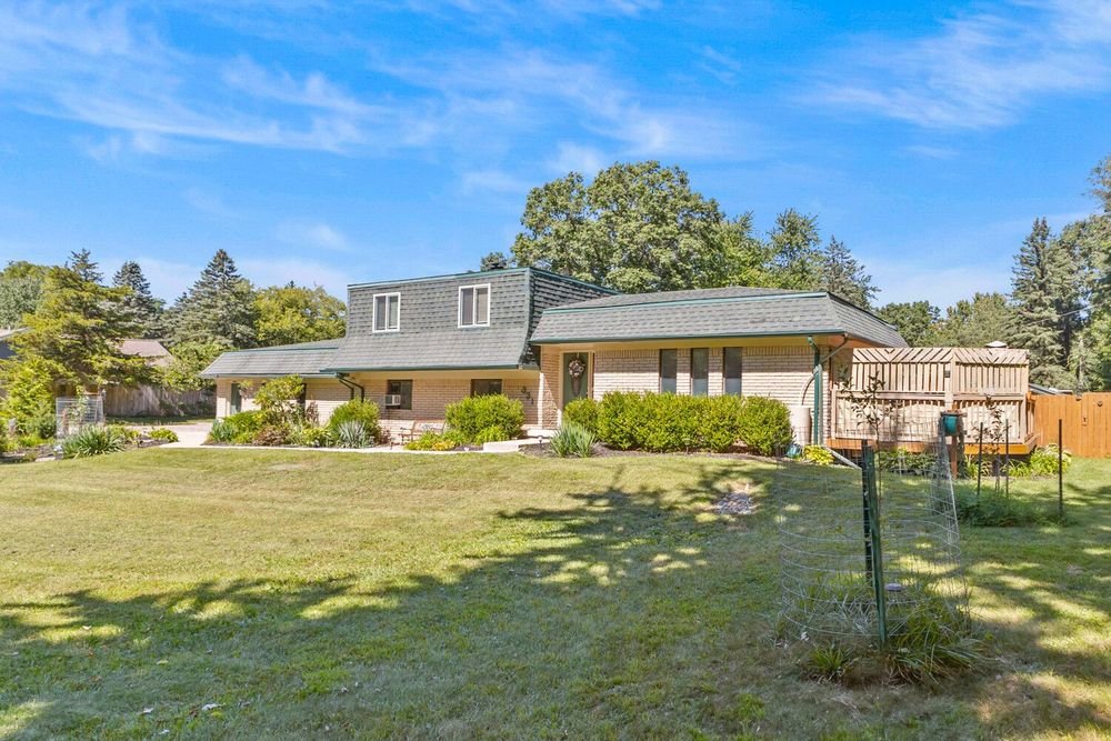 Ranch-style brick home with large green lawn, mature trees, and attached wooden fence on sunny day.