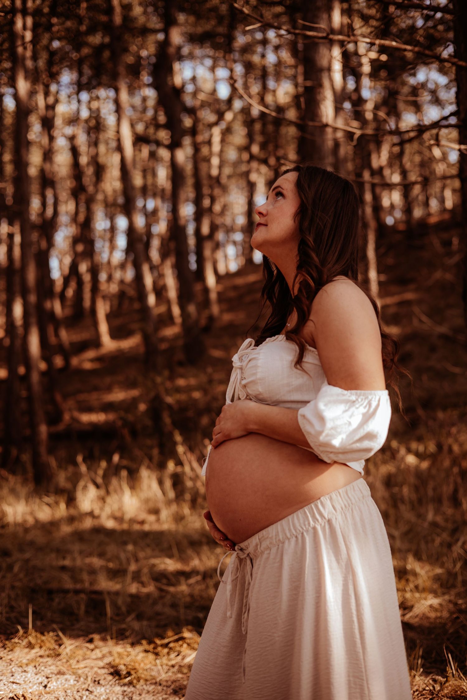 Pregnant woman in white off-shoulder dress holding baby bump while looking up in a sunlit pine forest.