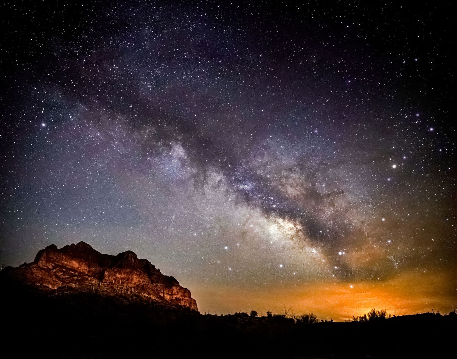 Stunning Milky Way galaxy arching over illuminated red rock mountains in a desert landscape at night.