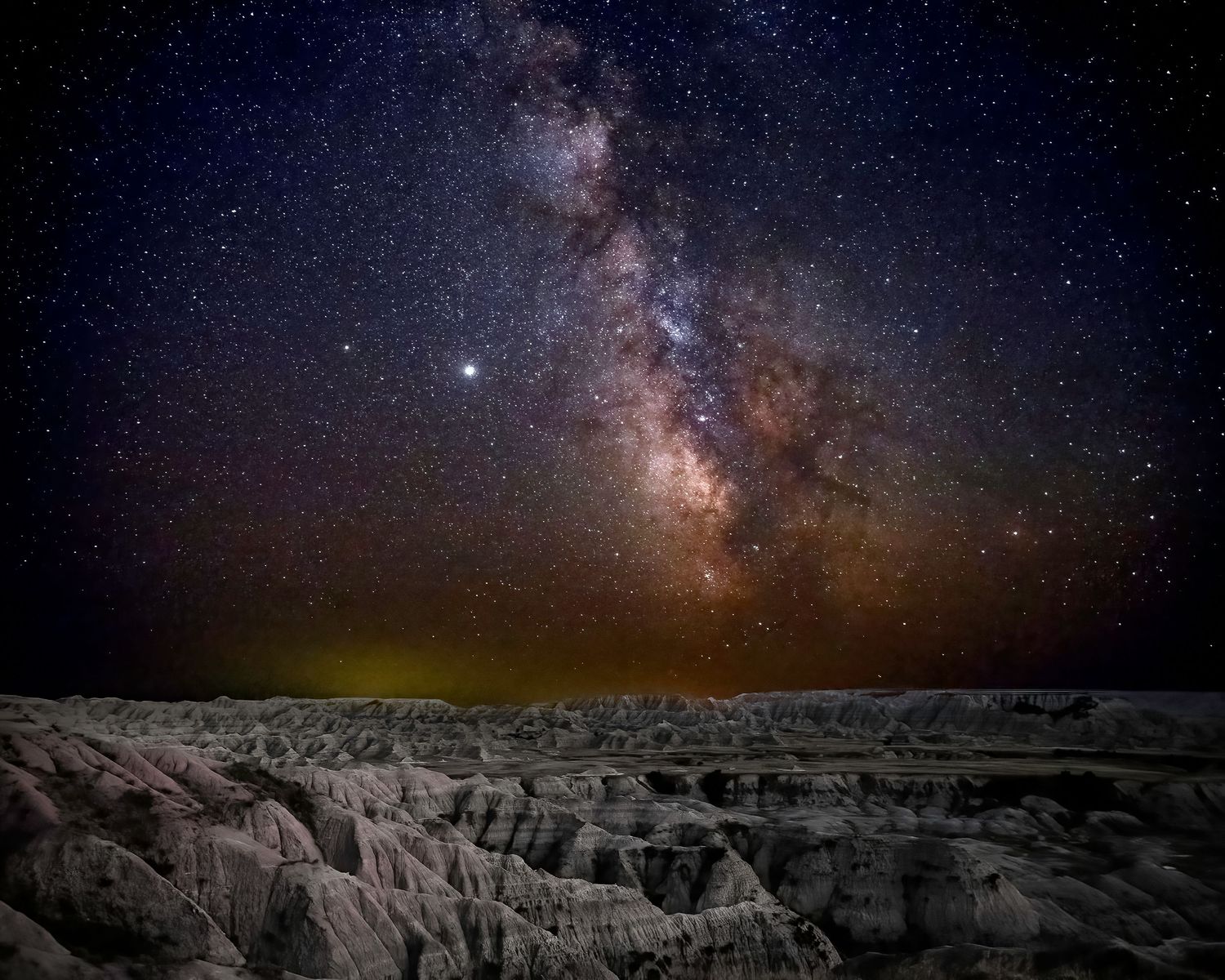Milky Way galaxy rising over dramatic eroded badlands rock formations under a star-filled night sky.