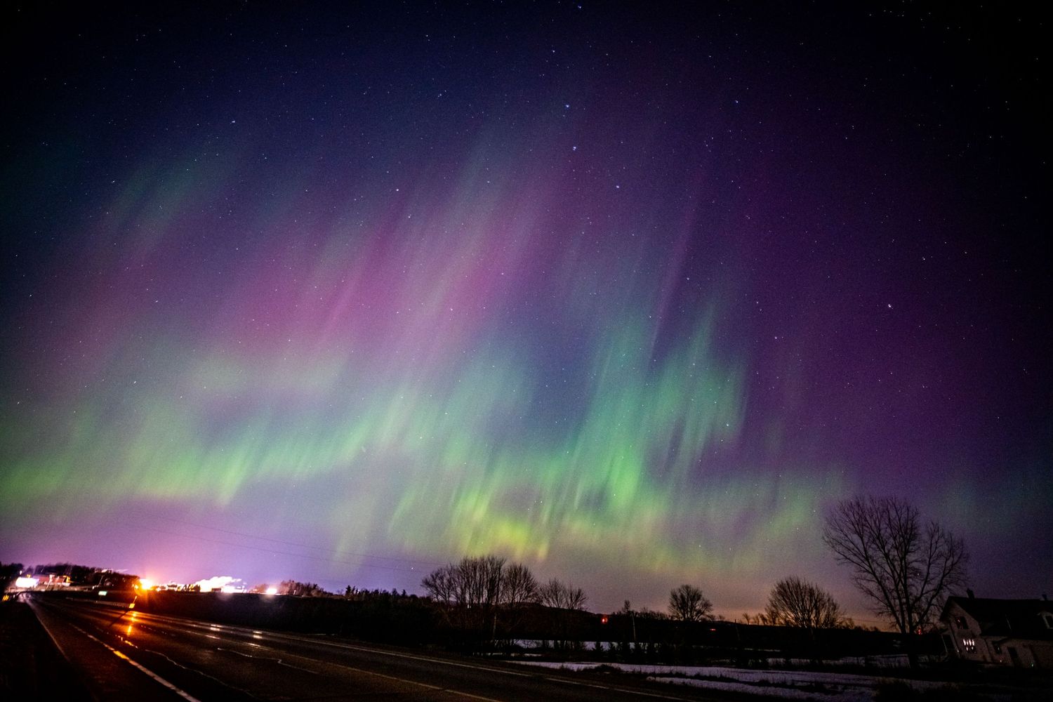 Vibrant aurora borealis with green and purple lights dancing across the night sky over a rural road.
