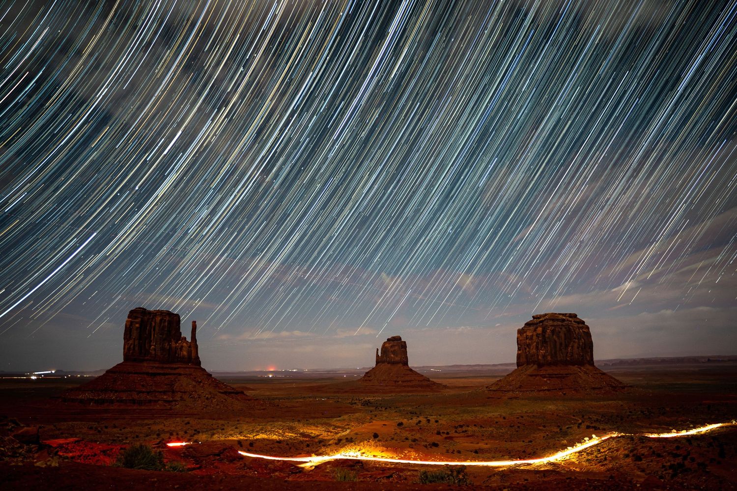 Long exposure star trails streak across the night sky above Monument Valley Mittens buttes with light trails.