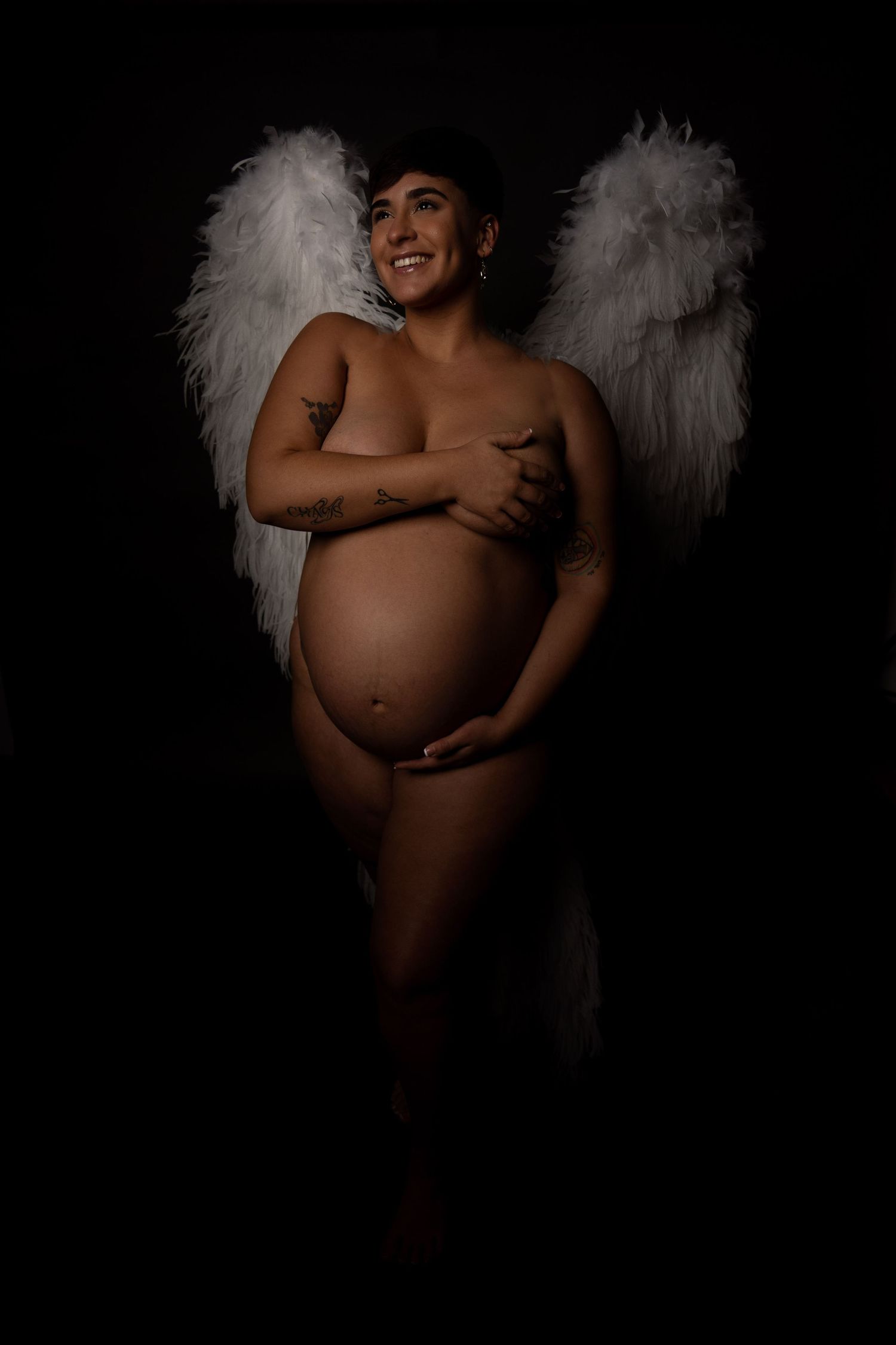 Pregnant woman with angel wings poses gracefully against dark background in artistic maternity photography.