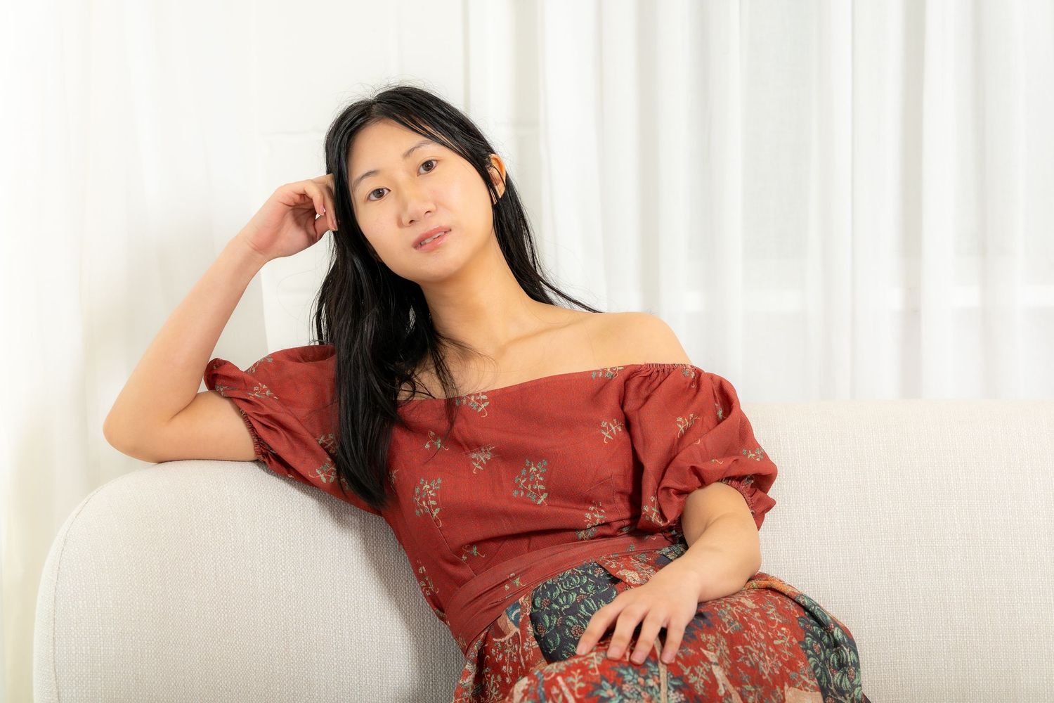 Young Asian woman in red floral off-shoulder dress sitting relaxed on white sofa.
