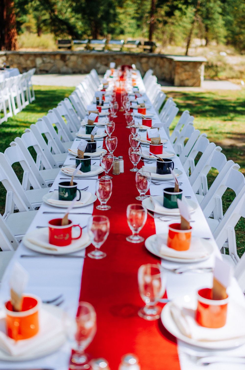 Long outdoor banquet table with red runner, white chairs, colorful mugs, and crystal glasses set for a formal event.