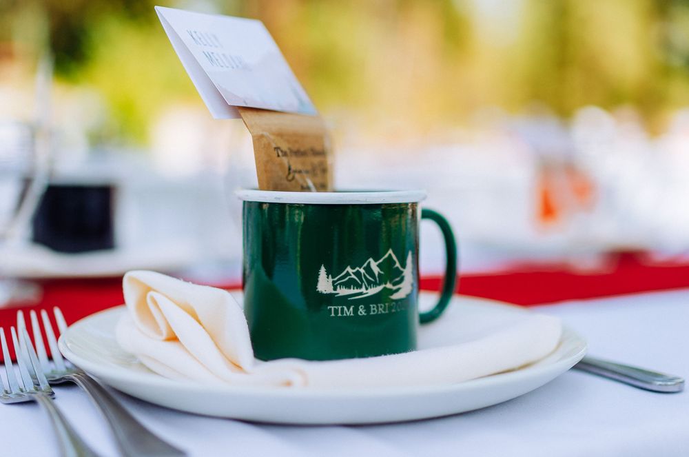 Green Tim & Bri wedding camp mug on white plate with napkin and silverware at outdoor reception table.