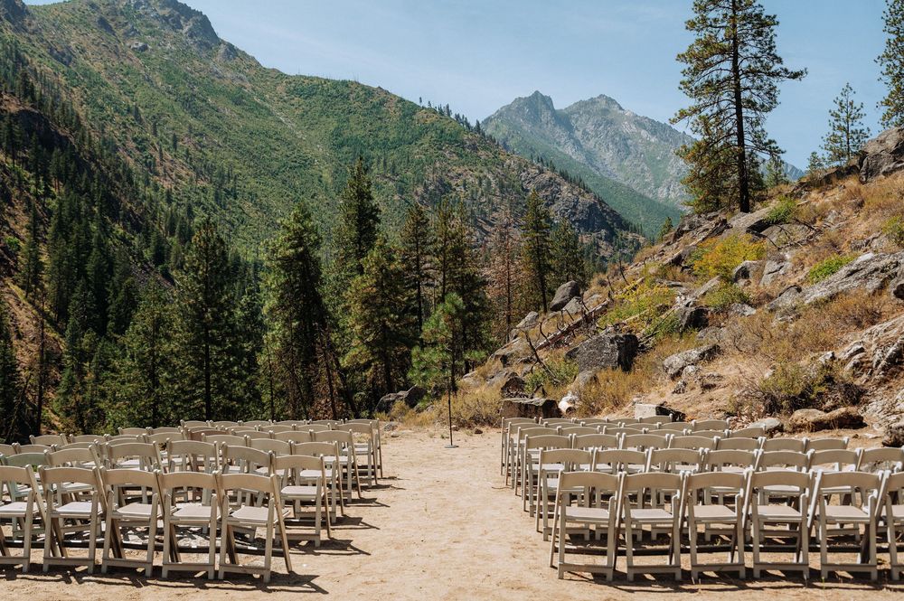 Outdoor wedding ceremony setup with white folding chairs arranged in rows against a scenic mountain and pine tree backdrop.