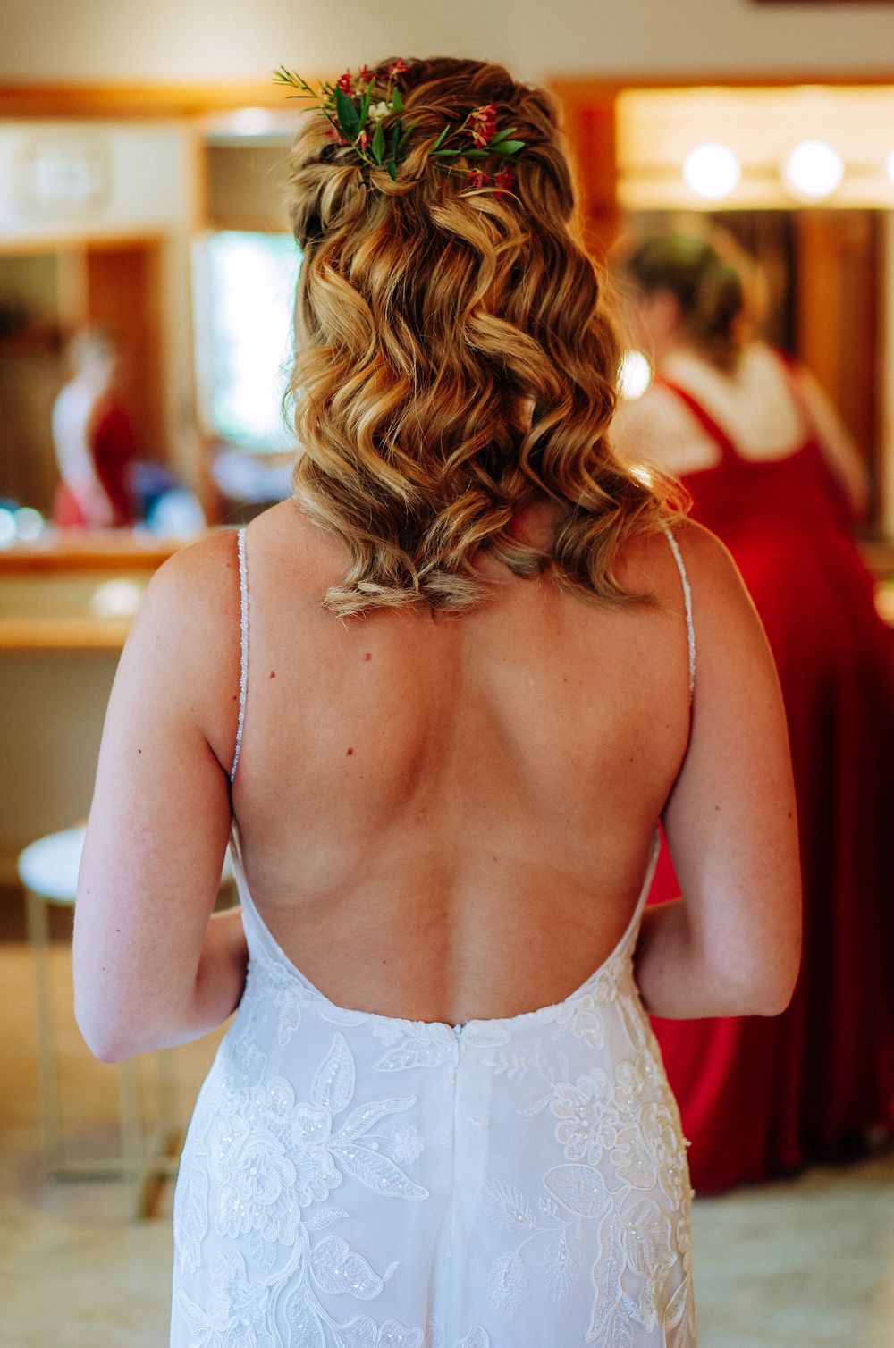 Bride in backless white lace gown with floral hair accessories, viewed from behind in a bridal suite.