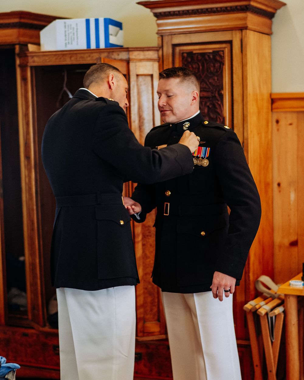 Two U.S. Marines in dress blues assist each other with uniform medals before a formal military ceremony.