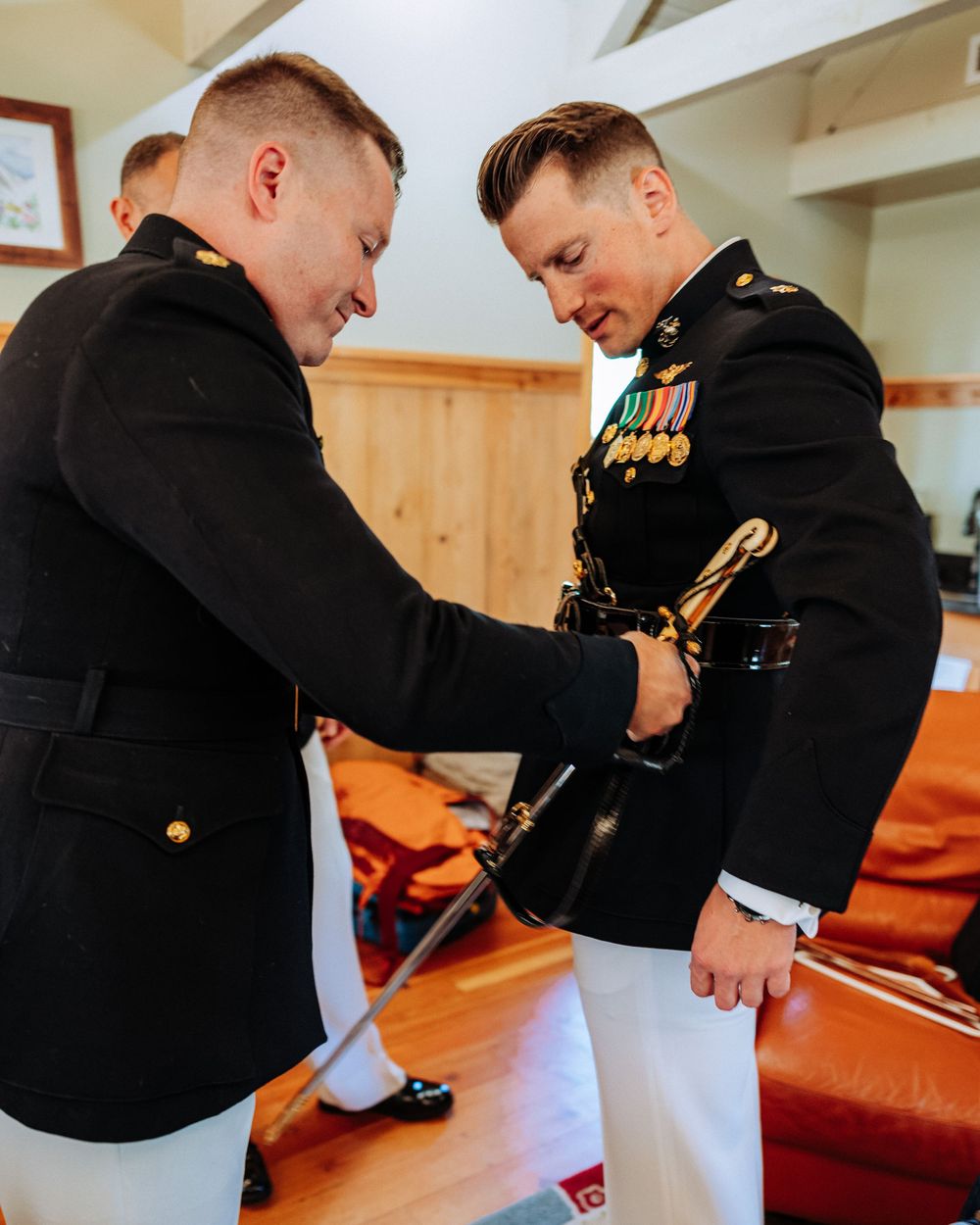 Two military officers in dress uniforms adjusting a ceremonial sword indoors.