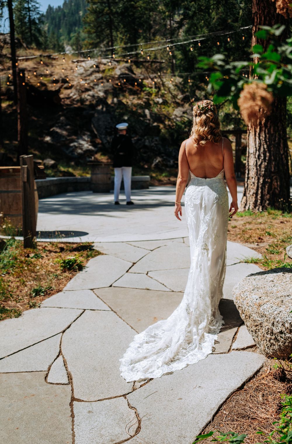 Bride in lace wedding gown with train walks toward groom in military uniform on stone path surrounded by pine trees.