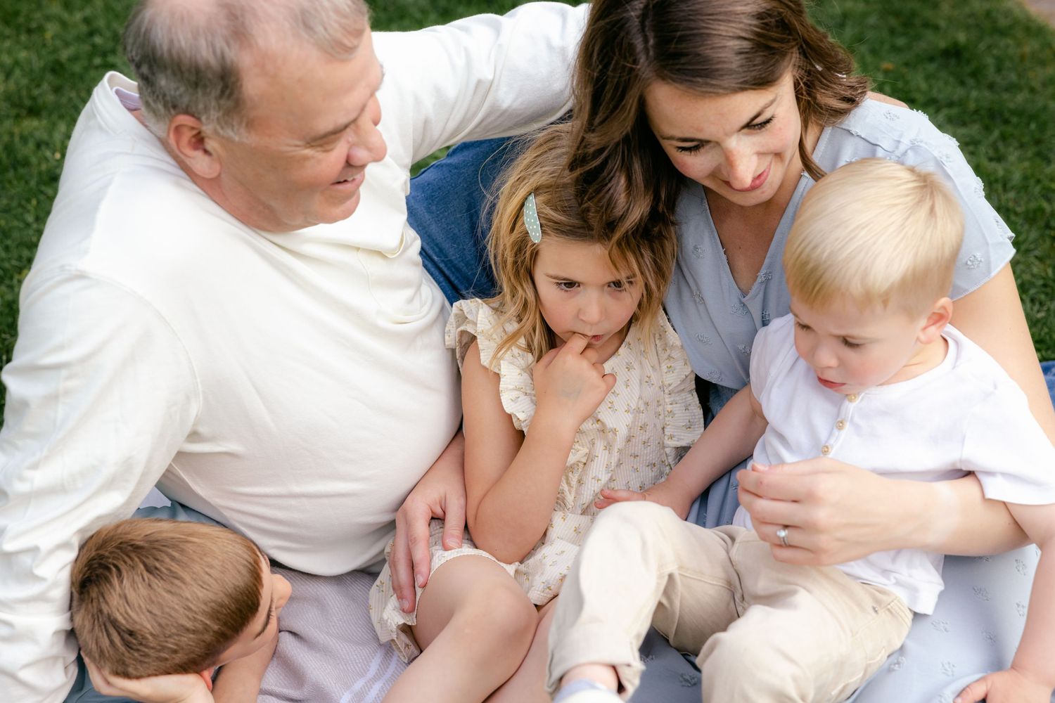 Family of five sitting on grass, parents and three young children sharing a warm, candid outdoor moment together.