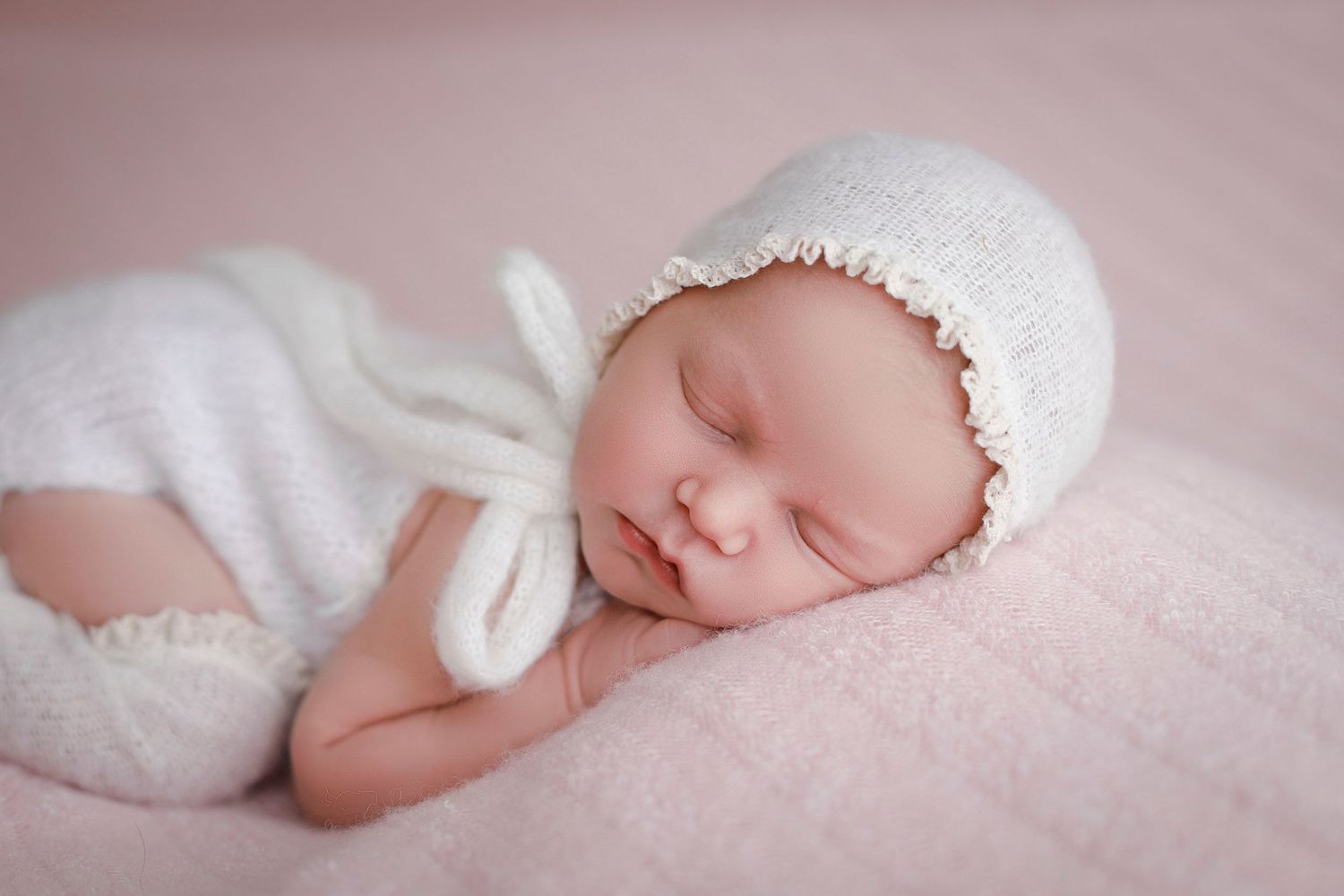 Newborn baby sleeping peacefully in white knit outfit and bonnet on soft pink blanket.