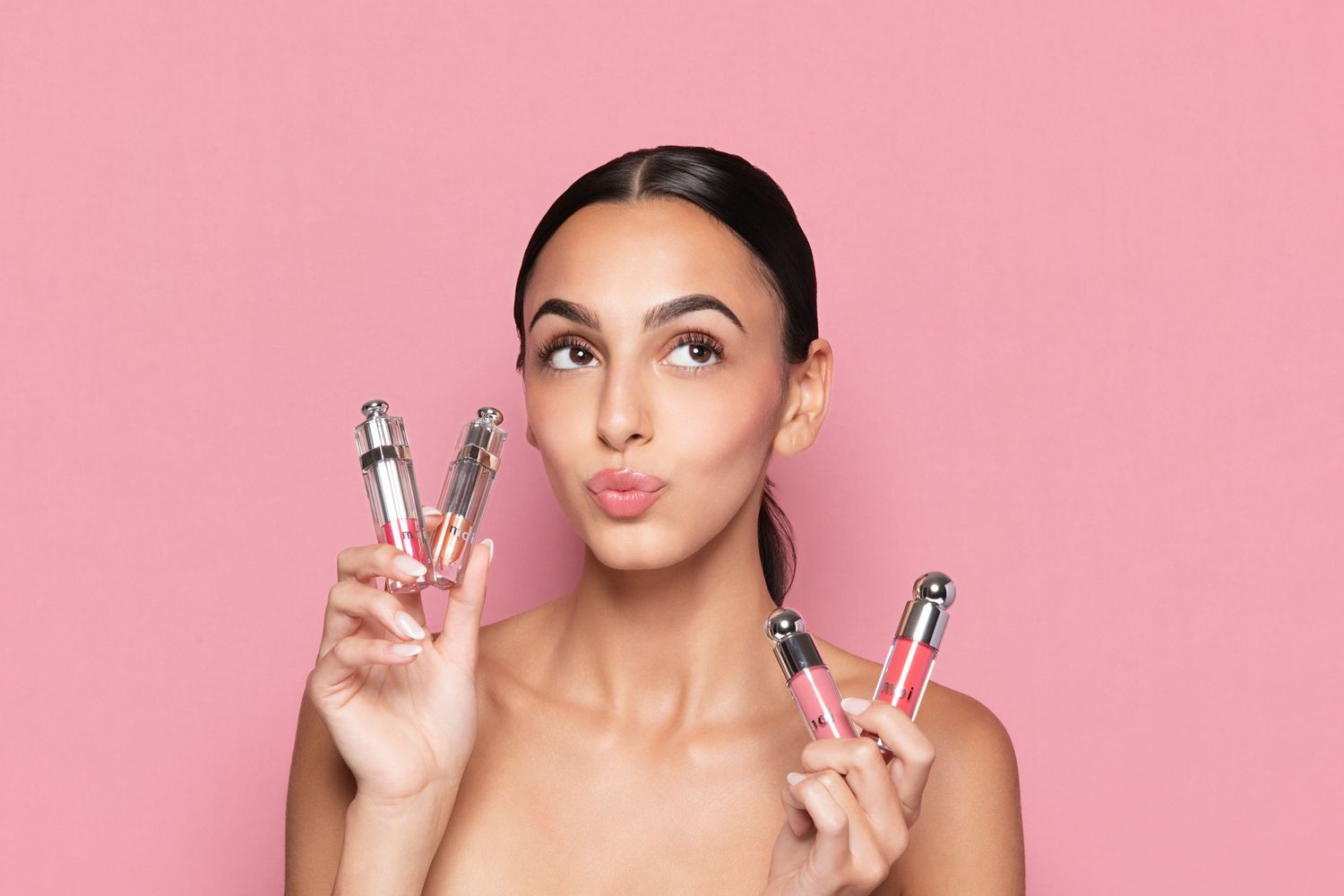 Woman holding multiple lip gloss tubes against a pink background, showcasing beauty products with a playful expression.