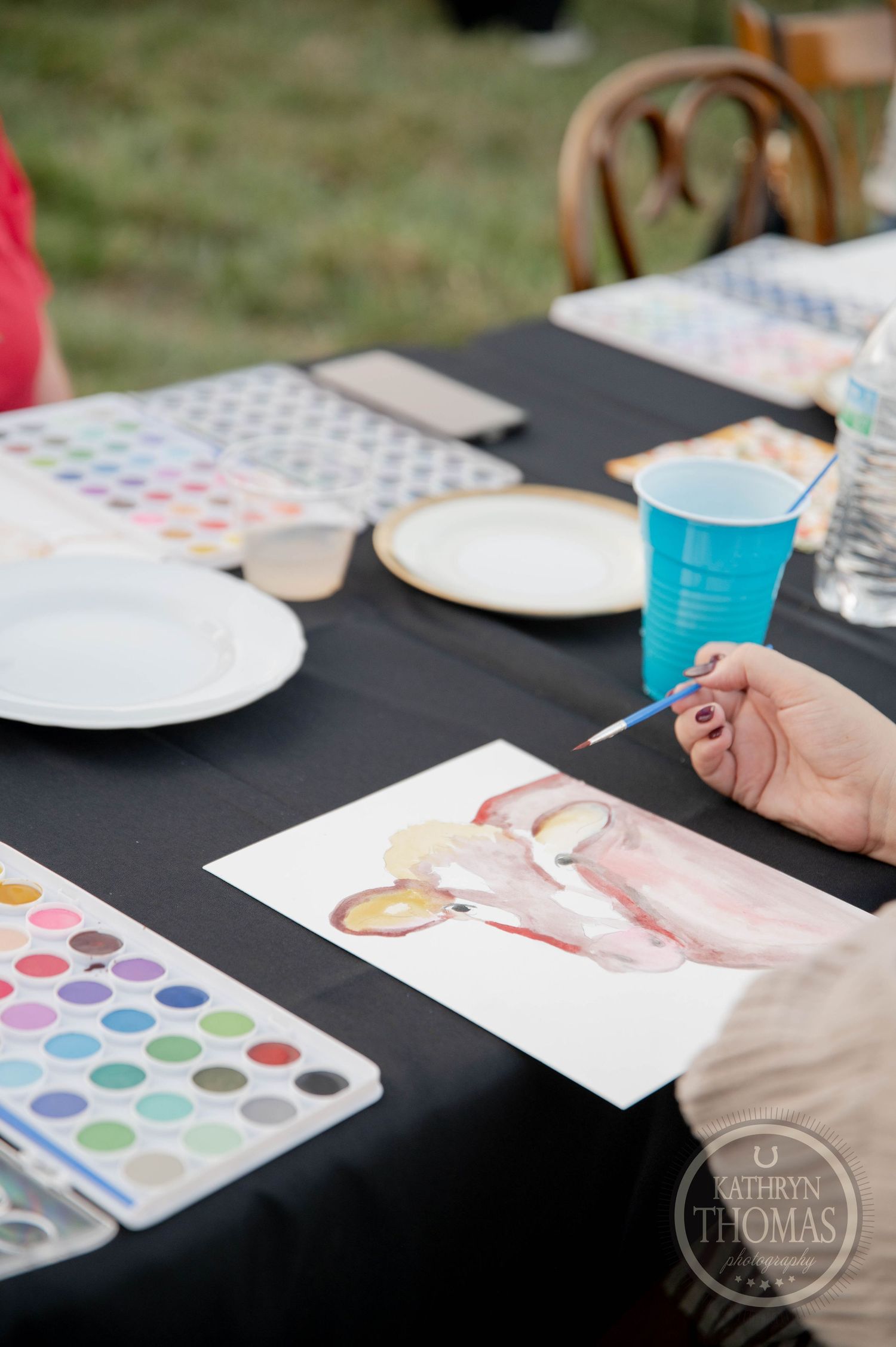 Person painting a watercolor cow at an outdoor art table with watercolor palette and blue cup nearby.