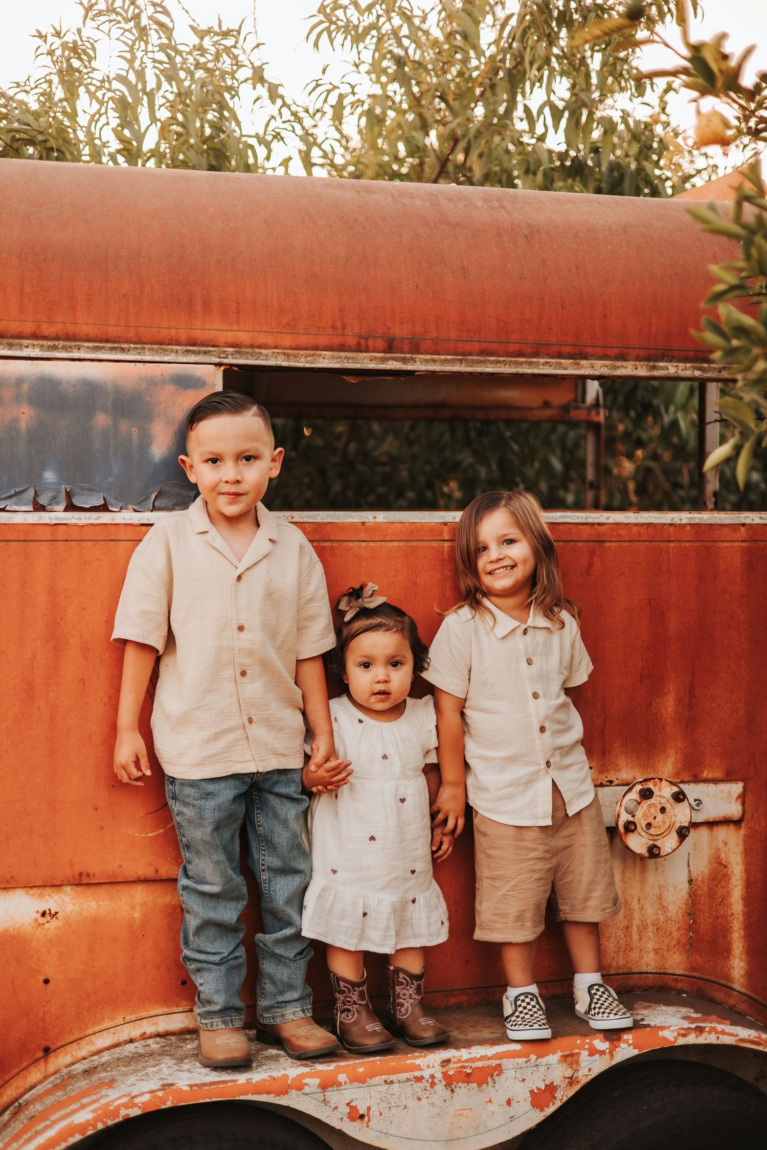 Three young children pose together in front of a rustic red vintage vehicle surrounded by greenery.
