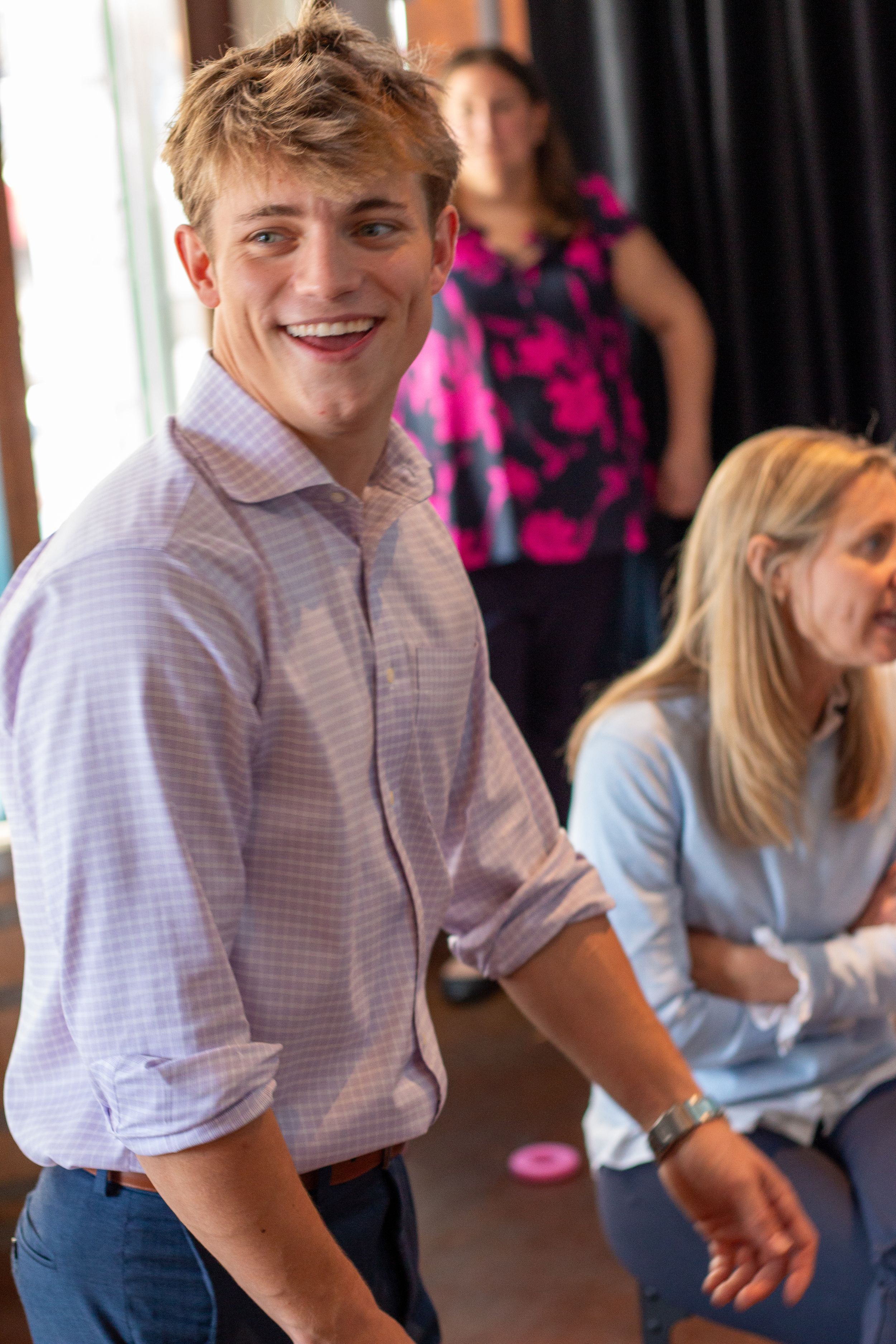Smiling young man in a light purple button-up shirt at an indoor event with women in the background.