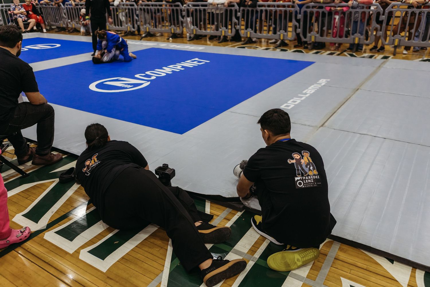 BJJ competitors and coaches sit courtside watching a match on blue competition mats at an indoor grappling tournament.