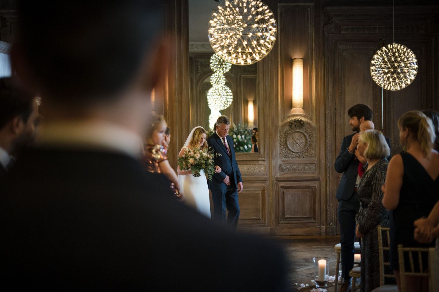 Elegant indoor wedding ceremony with glowing spherical chandeliers, guests standing, and couple at the altar in a wood-paneled room.