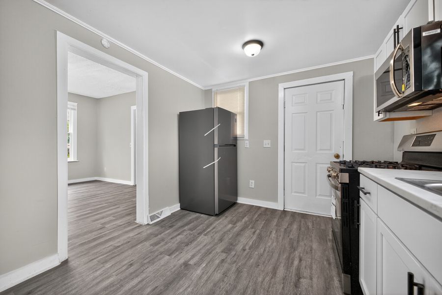Modern kitchen with stainless steel appliances, gray hardwood floors, white cabinets, and open doorway to adjacent room.