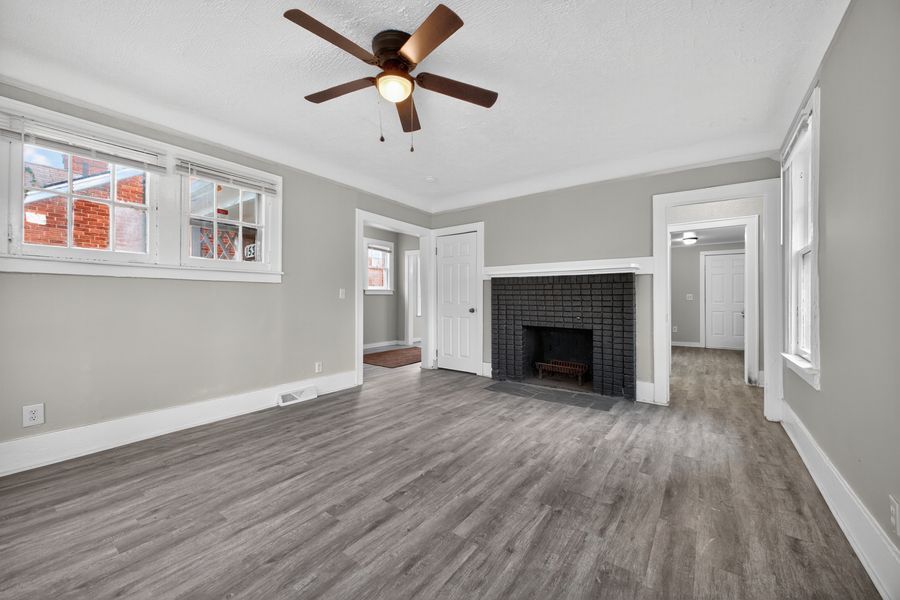 Bright living room with gray hardwood floors, brick fireplace, ceiling fan, and large windows with natural light.