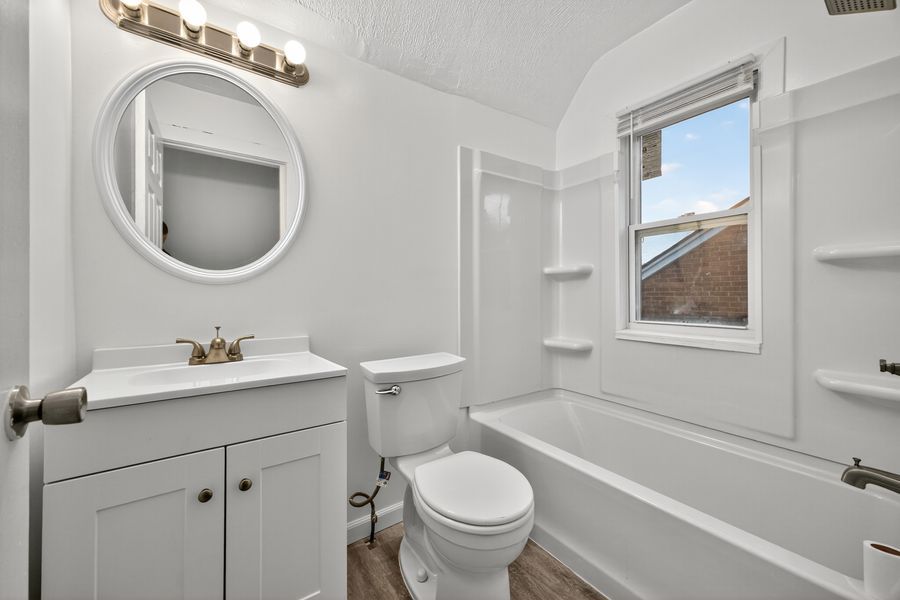 White bathroom with round mirror, single vanity, toilet, and bathtub-shower combo with a window above.