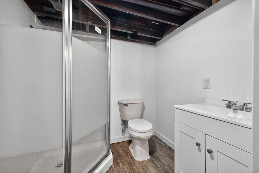 Basement bathroom with glass shower, white toilet, vanity, wood-look floors, and exposed black ceiling beams.