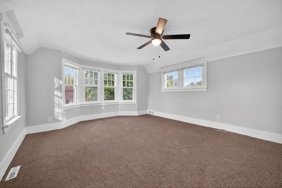 Spacious bedroom with brown carpet, ceiling fan, bay window, and abundant natural light.
