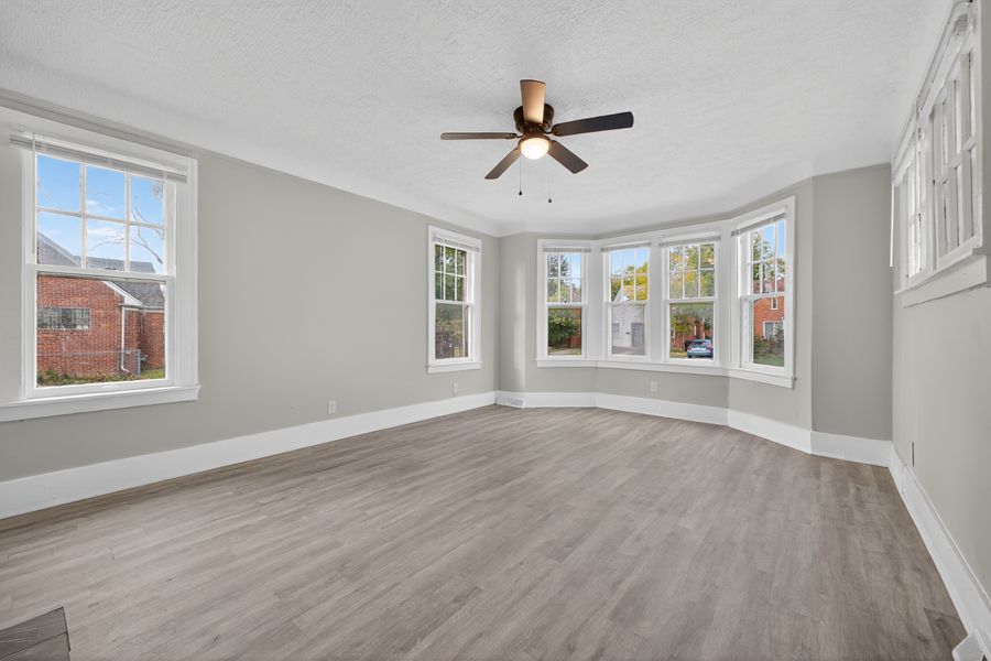 Bright empty living room with hardwood floors, ceiling fan, bay windows, and fresh gray paint on textured walls.