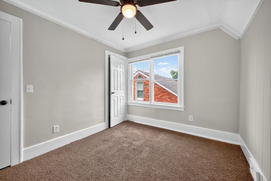 Bright bedroom with brown carpet, crown molding, ceiling fan, and a window overlooking a red brick building.