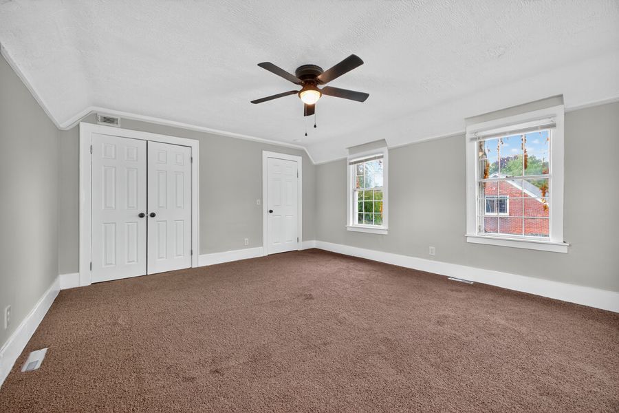 Spacious bedroom with brown carpet, crown molding, ceiling fan, white closet doors, and two windows with natural light.
