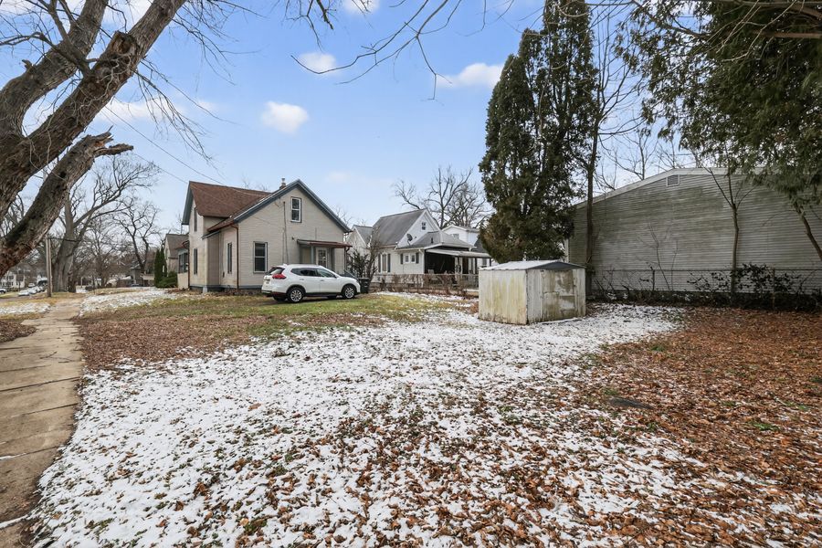 Snow-dusted backyard of a residential property with a white SUV parked beside a charming house and a storage shed.