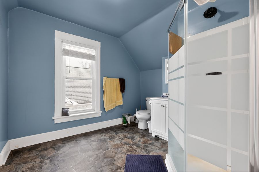 Blue bathroom with vaulted ceiling, walk-in shower, white vanity, toilet, and window with yellow towel.