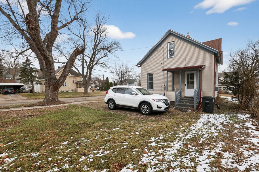 White SUV parked in front of a small beige home with a large tree and light snow on the ground.