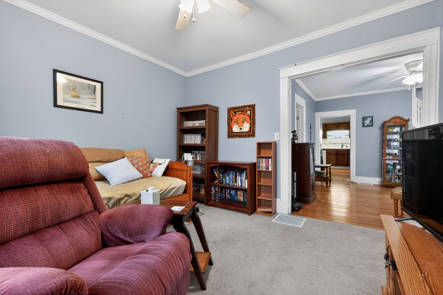 Cozy living room with red recliner, carpet, crown molding, ceiling fan, and bookcase visible through open doorway.