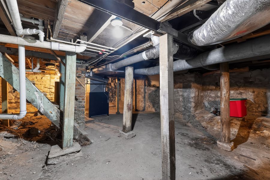 Unfinished basement with exposed pipes, ductwork, stone walls, wooden support columns, and concrete floor.