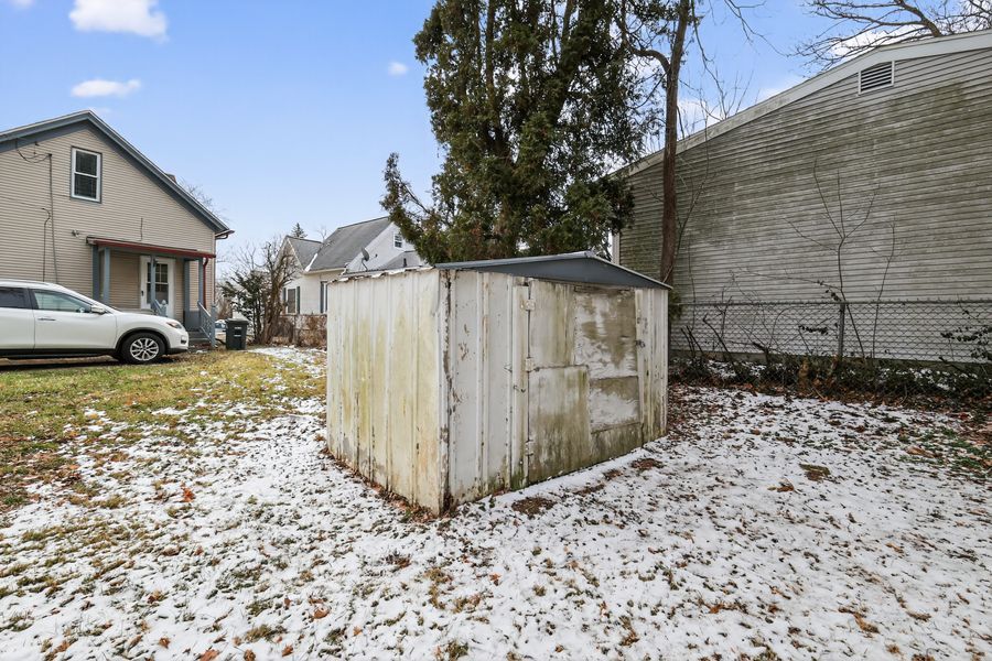 Small metal storage shed in backyard with light snow on ground, surrounded by trees and neighboring homes.