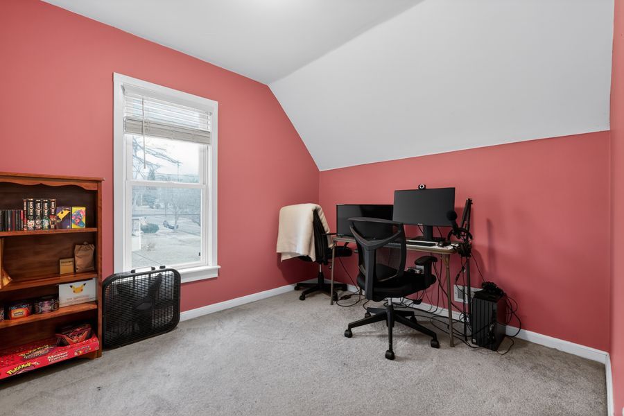 Pink-walled home office with sloped ceiling, computer desk, ergonomic chair, and carpeted floor near a bright window.