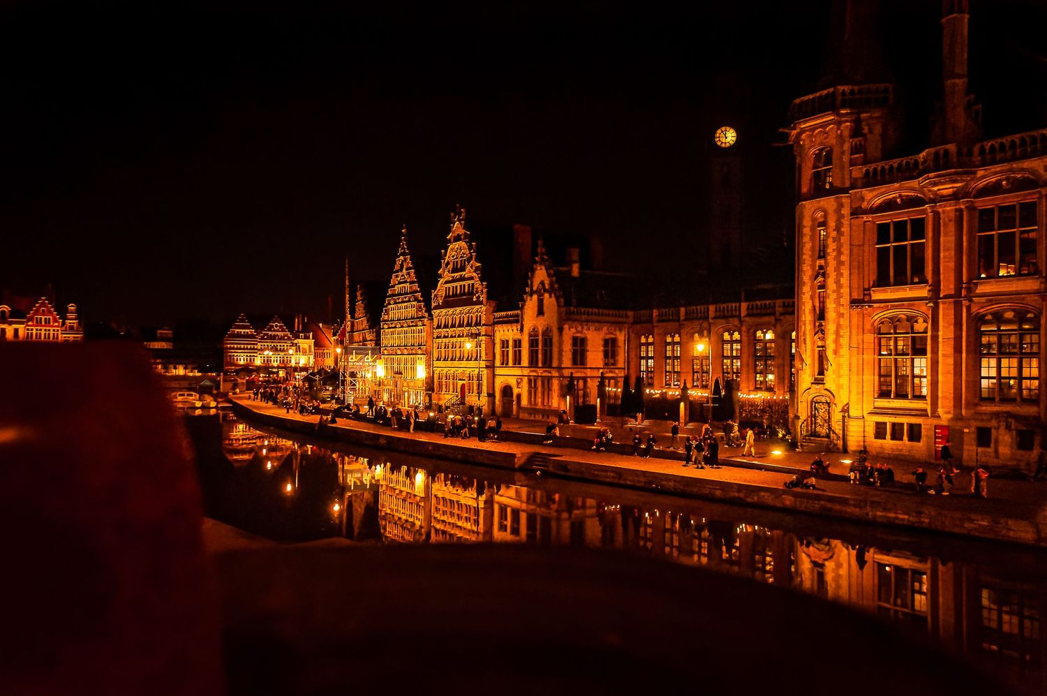 Wide nighttime photo of a brightly lit city square with historic buildings and street lights, reflecting off wet pavement. Gent Belgium