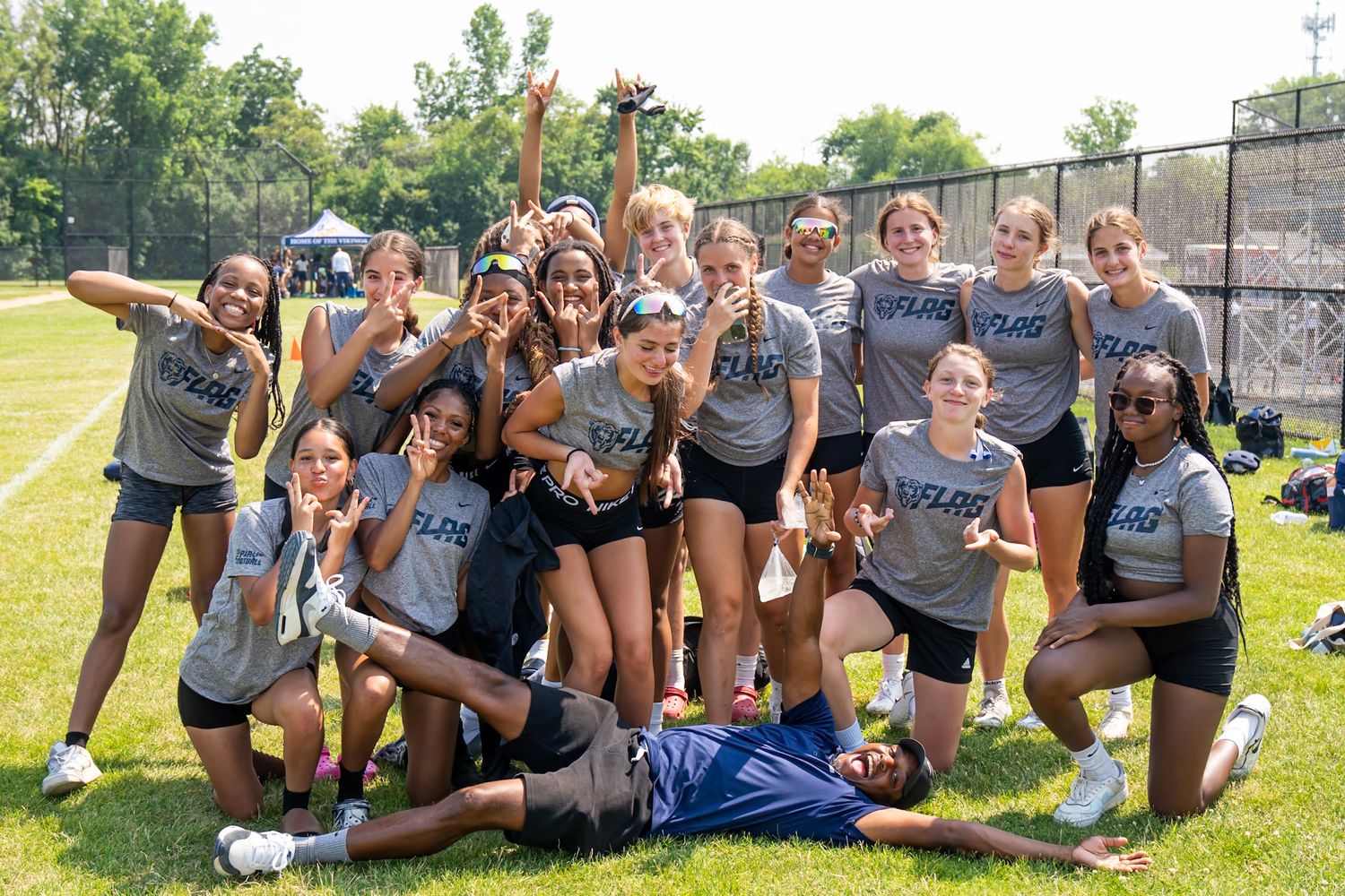 A girls sports team poses together on a green field, wearing matching gray shirts, celebrating with smiles and fun poses.