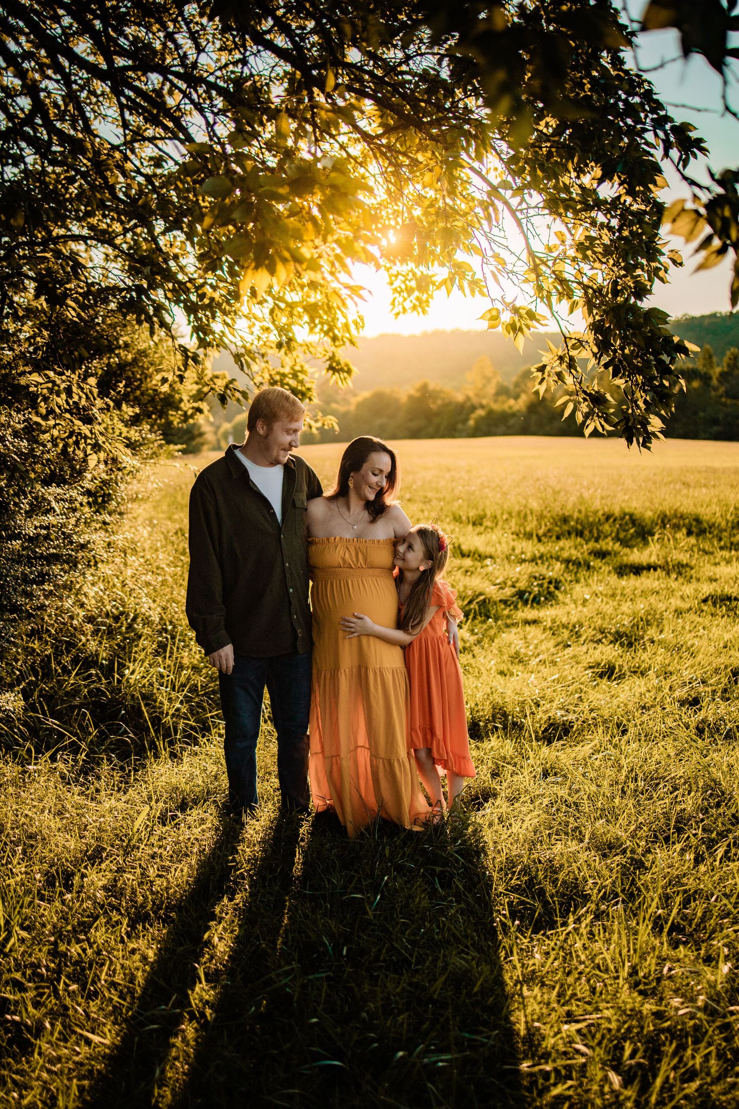 A family poses at golden hour under a tree, with a pregnant woman in a yellow dress, her partner, and young daughter in orange.