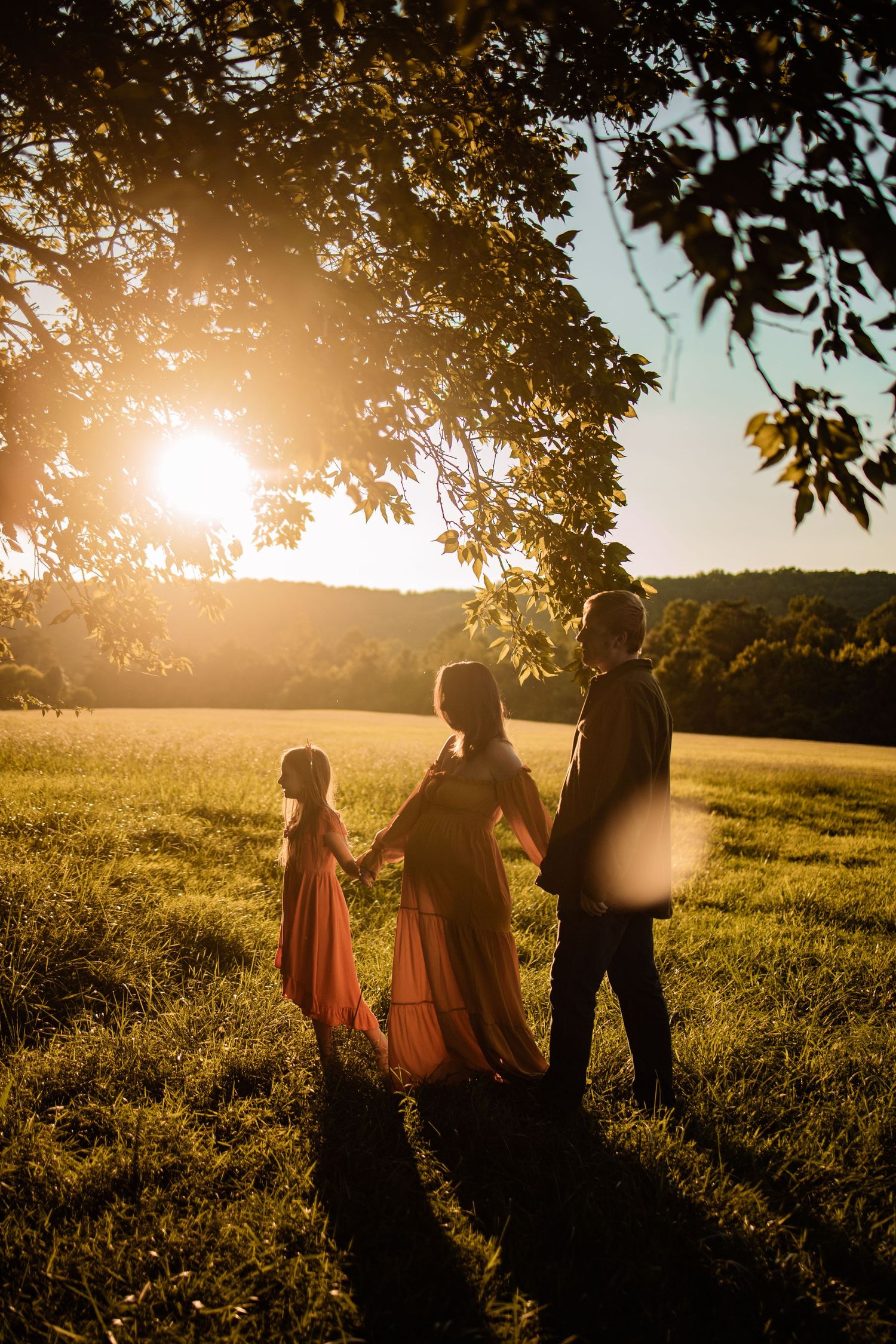 Family of three holding hands at golden sunset in a green field, woman in flowing red dress with young girl.