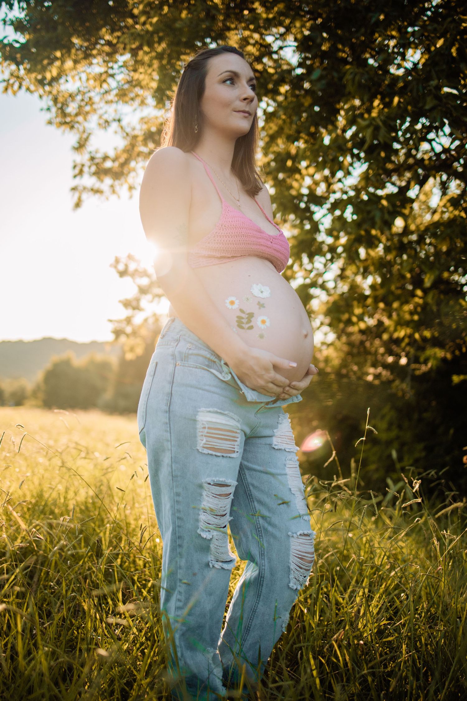 Pregnant woman in pink crop top and ripped jeans holding her belly outdoors at golden sunset.