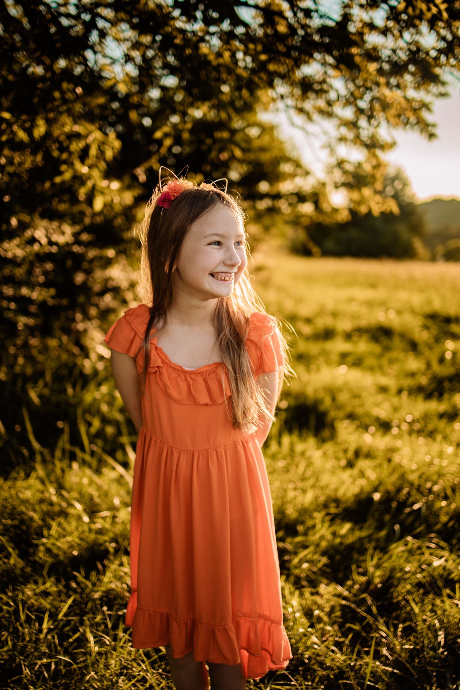A smiling young girl in an orange dress with cat ears headband stands in a sunlit green field at golden hour.