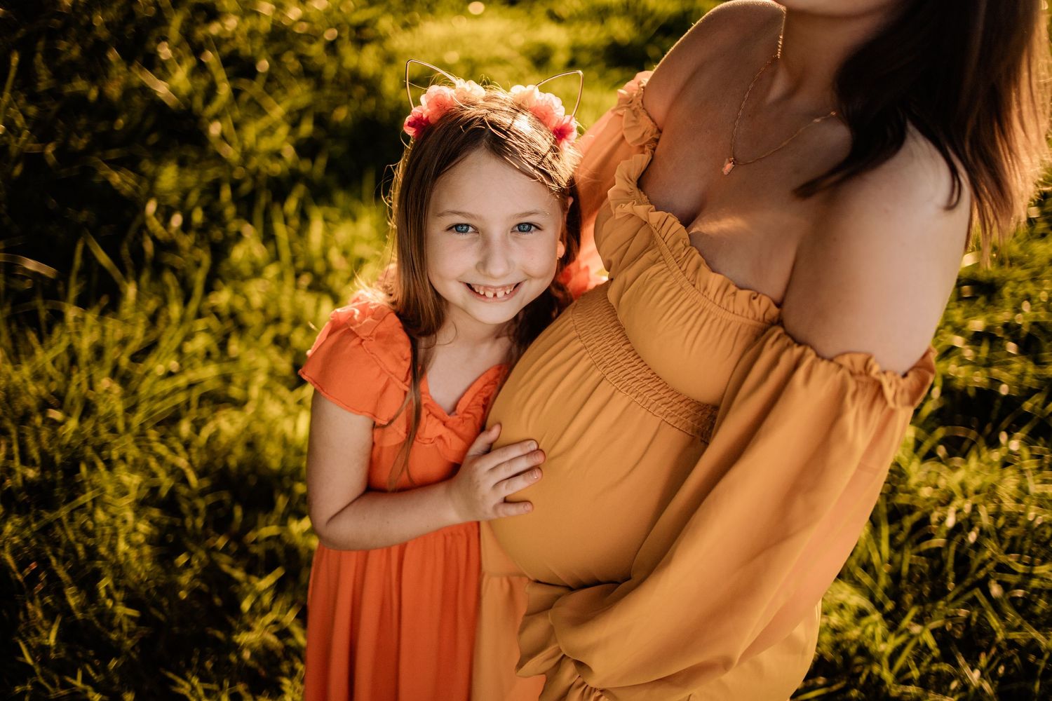 A young girl in an orange dress smiles beside a pregnant woman in a golden dress outdoors in sunlight.