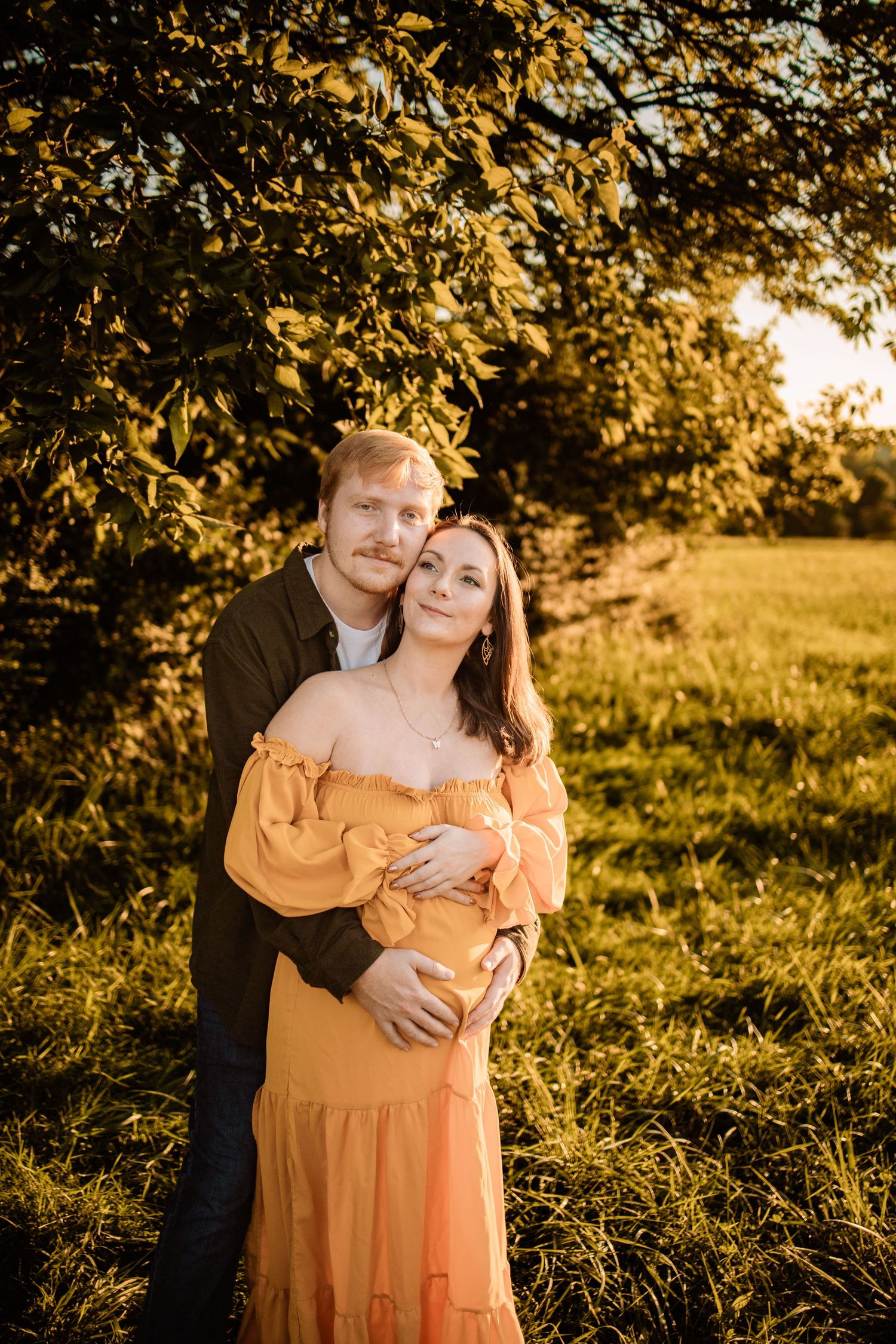 Couple embracing at golden hour sunset near lush green trees, woman in orange off-shoulder dress, man in dark shirt.