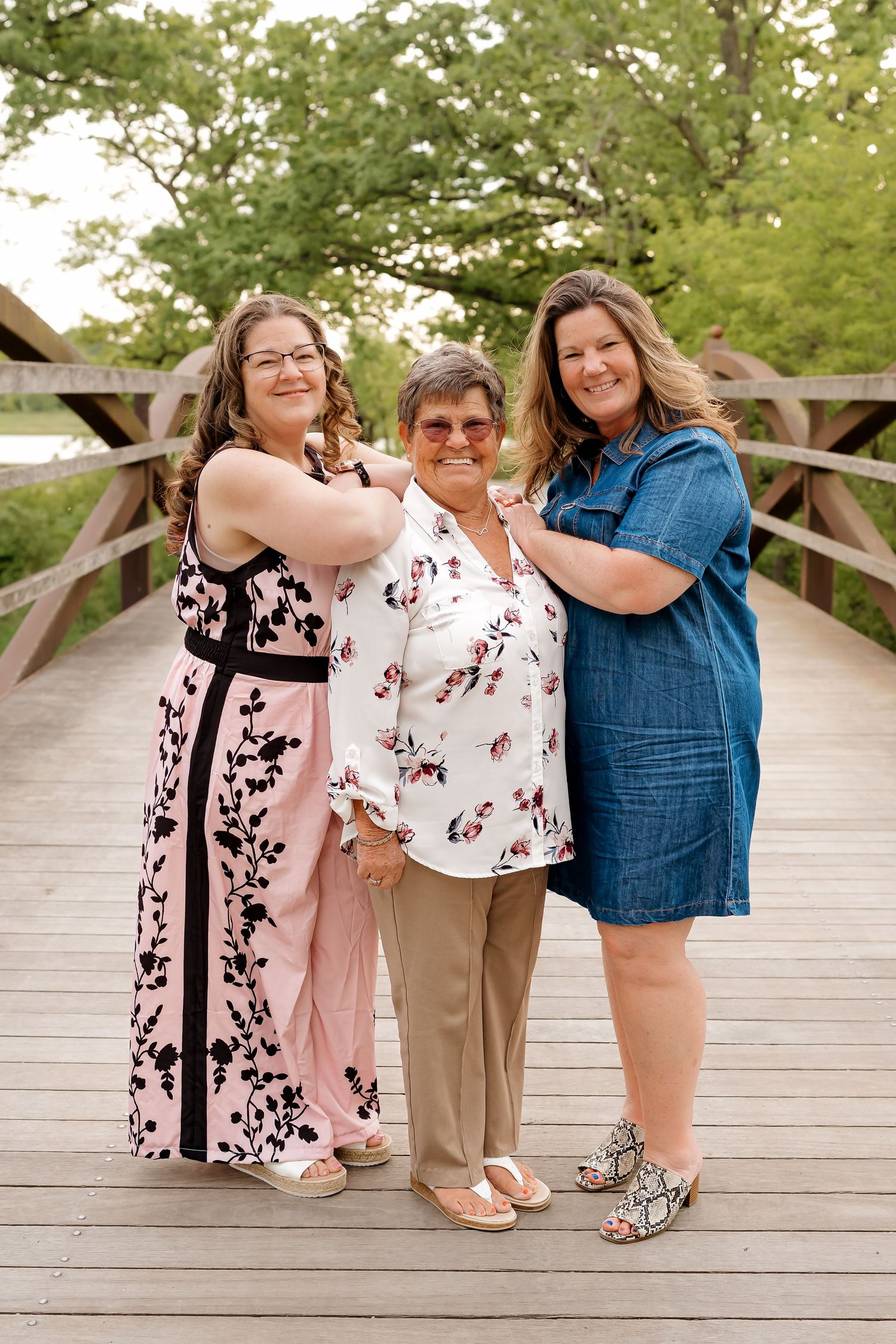 Three women smiling together on a wooden bridge surrounded by green trees, posing for a family portrait outdoors.