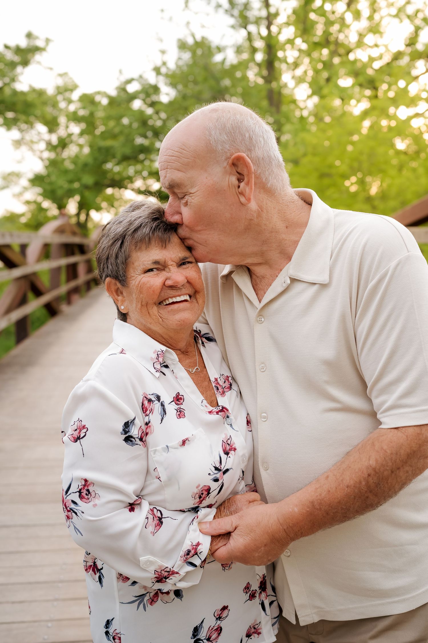 An elderly couple shares a tender moment on a wooden bridge, as the man kisses the smiling woman on the cheek.