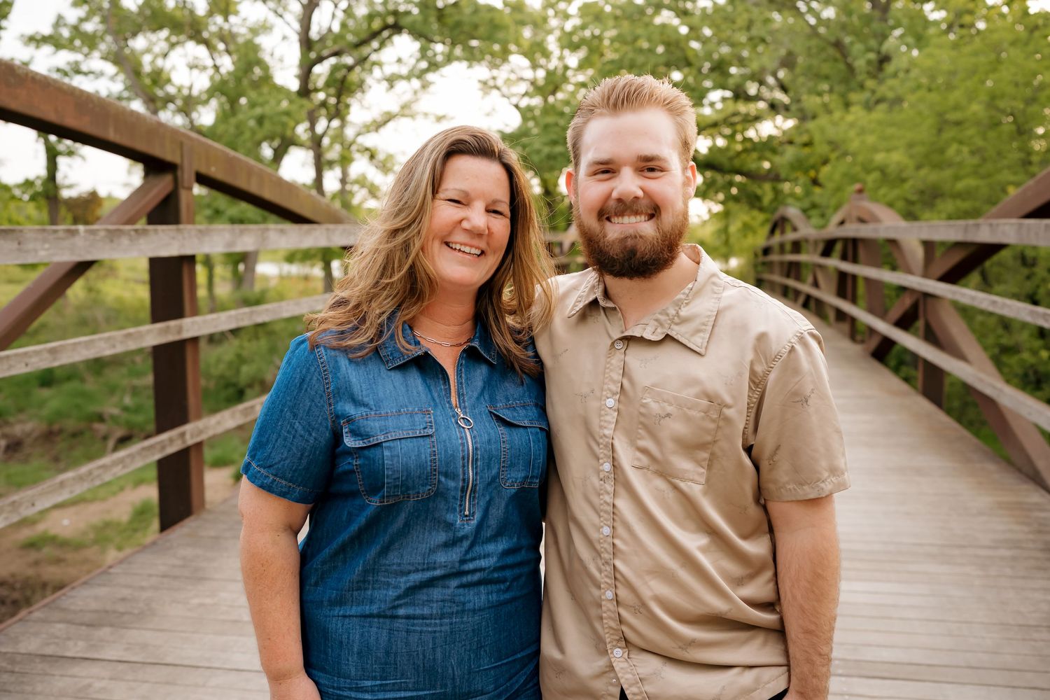 A smiling woman in a blue dress and a bearded man in a tan shirt pose together on a wooden bridge outdoors.